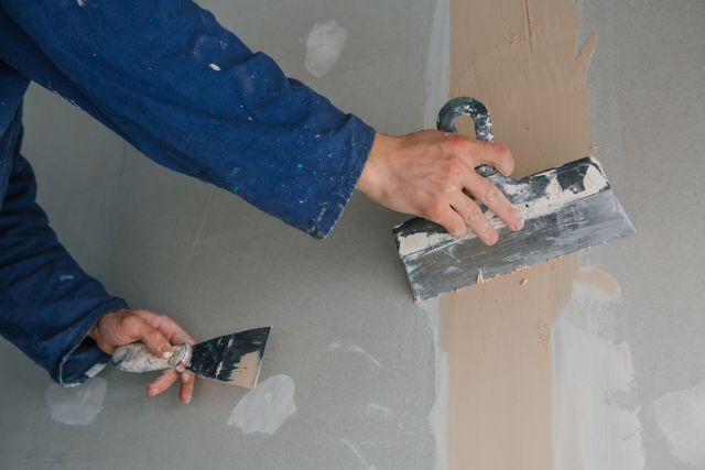 Plasterer man works plastering two trowels on plasterboard in blue uniform